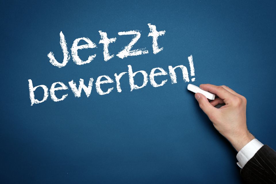 Eine Hand schreibt mit Kreide "Jetzt bewerben!" an eine Tafel