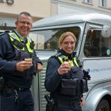 Ein Polizist und eine Polizistin trinken vor einem historischen Kaffee-Mobil einen Kaffee.