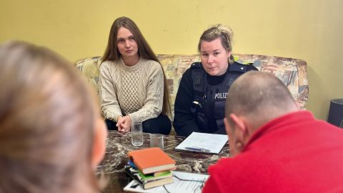 A policewoman and a member of the emergency counseling team are sitting in the living room of the relatives of a fatality. They look serious. The relatives are sitting opposite them. They can be seen from behind in the foreground.