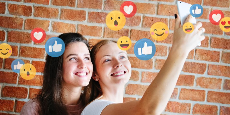 Zwei junge Frauen machen zusammen ein Selfie. Über ihnen sind Social Media Icons wie Emojis Herzen und Daumen hoch zu sehen. Im Hintergrund ist eine Backsteinmauer zu sehen.